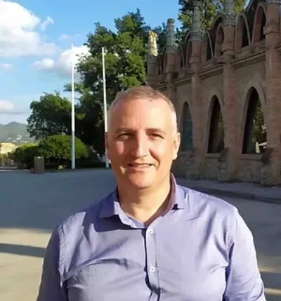Hombre sonriendo al aire libre con una camisa lila junto a un edificio de piedra y árboles en un día soleado.