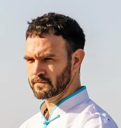 Hombre con camisa blanca con ribete turquesa, mirando hacia la izquierda al aire libre bajo un cielo despejado.