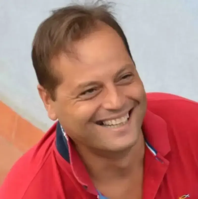 Hombre sonriente con camisa roja mirando hacia un lado, sobre un fondo claro.