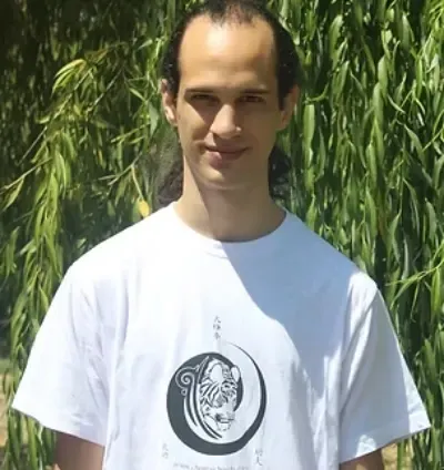 Persona con camiseta blanca de pie al aire libre frente a follaje verde.