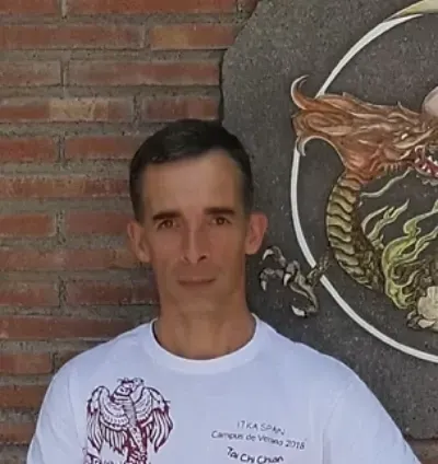 Un hombre, de pie junto a una pared de ladrillos, al lado de un mural circular con un dragón, viste una camiseta blanca.
