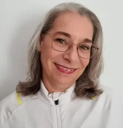 Persona sonriente con cabello gris y gafas, vestida con una blusa blanca, sobre un fondo liso.