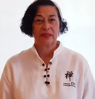Mujer con camisa blanca de botones negros, de pie frente a un fondo liso, mirando a la cámara.