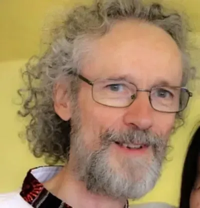 Hombre sonriente con cabello gris rizado, gafas y barba sobre un fondo amarillo.