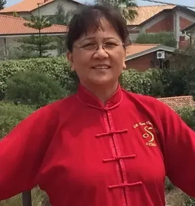 Persona sonriente con una chaqueta roja tradicional de pie al aire libre con casas y vegetación detrás.