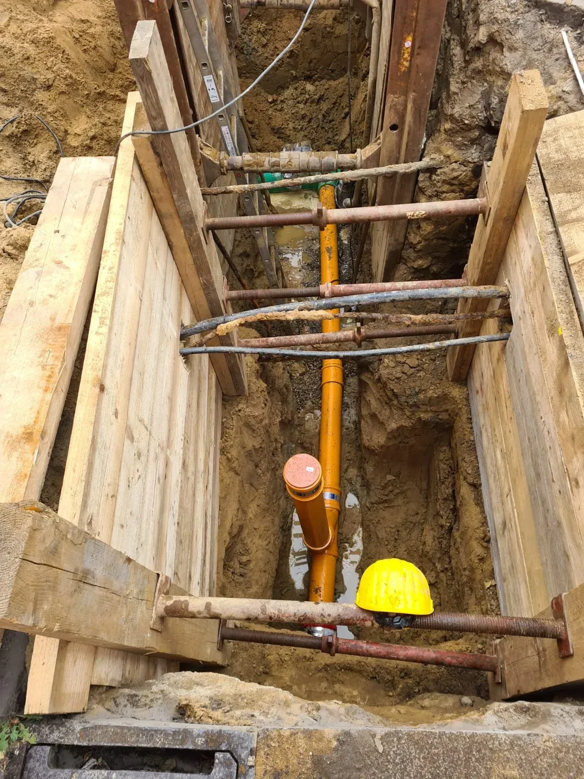 Graben mit Holzstützen, in dem sich ein orangefarbenes Rohr, Kabel und ein Schutzhelm befinden.