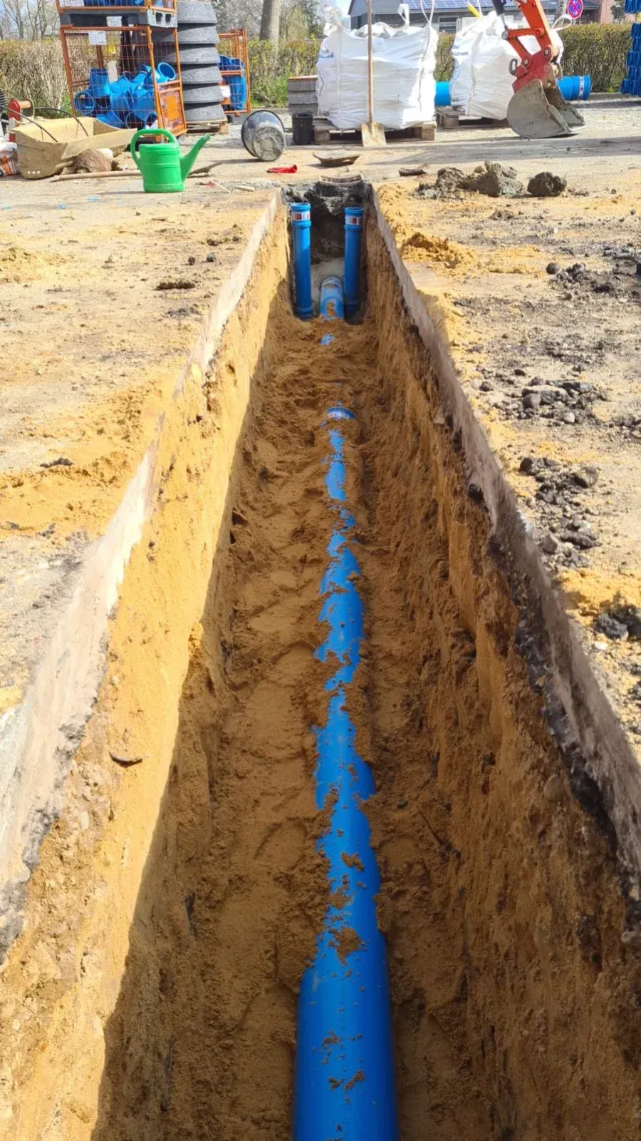 Blaues Wasserrohr in einem Graben, Baustelle mit Sand, Werkzeugen und Material im Hintergrund.