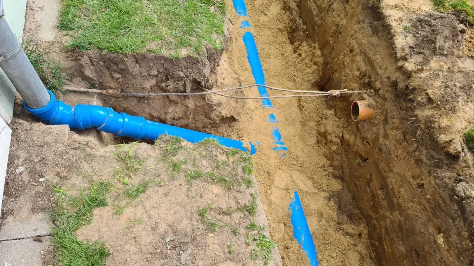 Blaues Abflussrohr in einem Graben, angeschlossen an ein Fallrohr, inmitten von Erde und Gras.