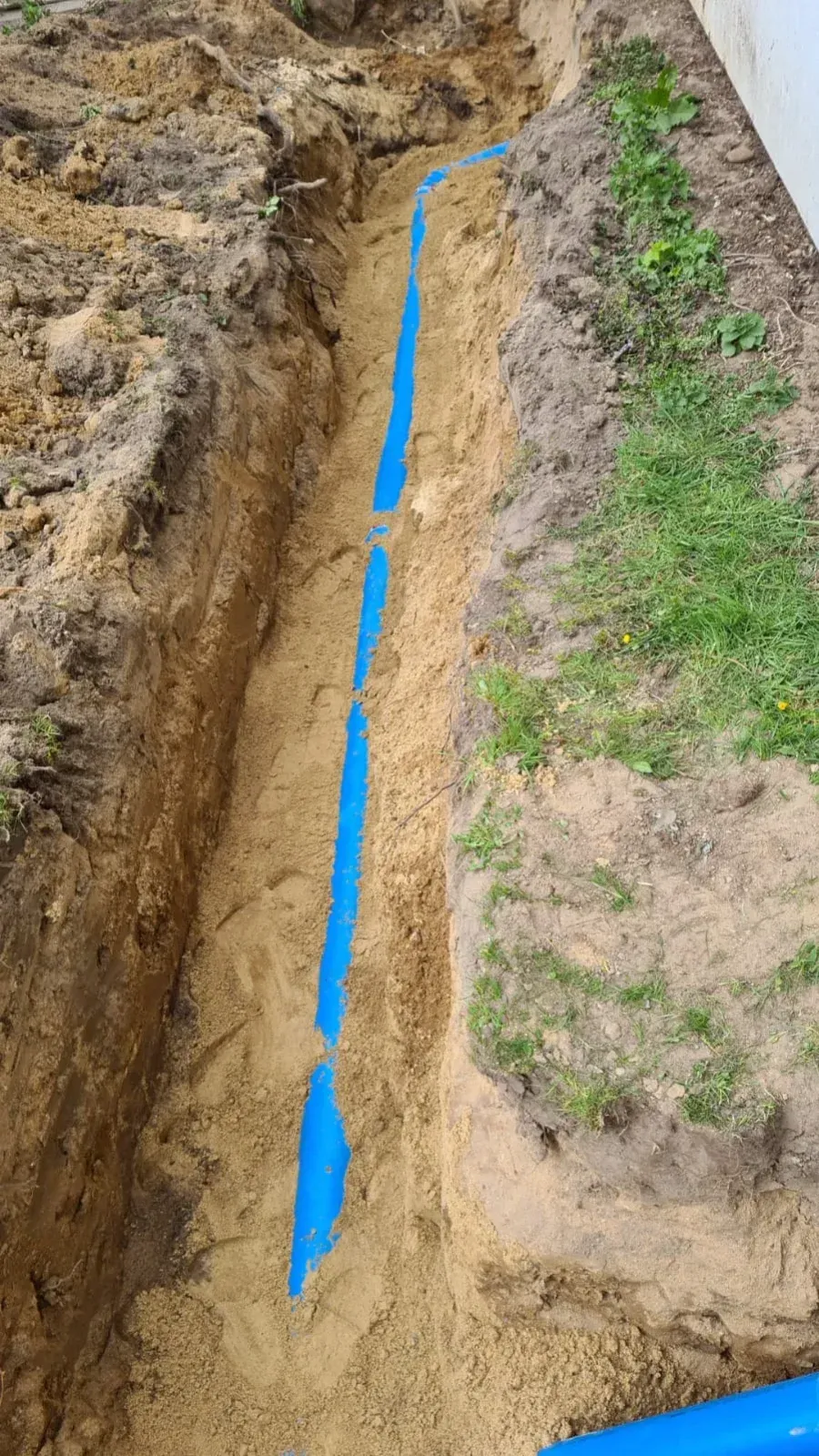 Ein in den Boden gegrabener Graben, an dem blaue Rohre entlanglaufen, in der Nähe von Gras und einer Mauer.