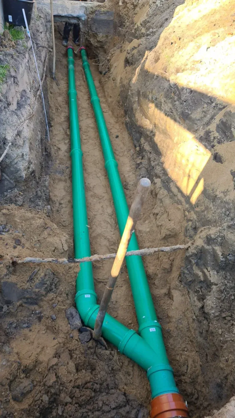 Grüne Abflussrohre in einem Graben vereinigen sich zu einem braunen Rohr. Eine Schaufel liegt quer über den Rohren.