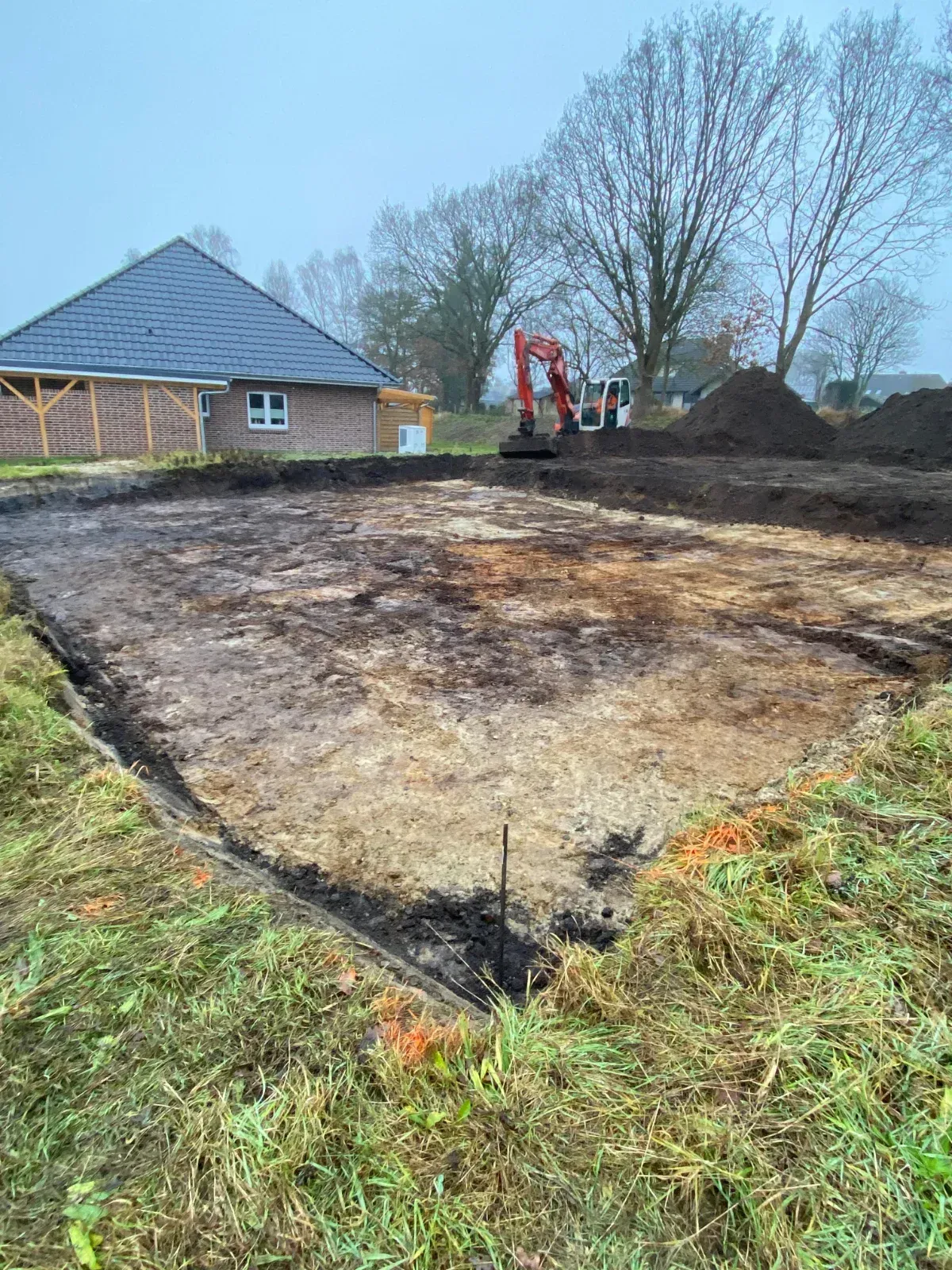 Ausgehobene Baustelle mit einem Bagger im Hintergrund; ein Haus ist ebenfalls zu sehen.
