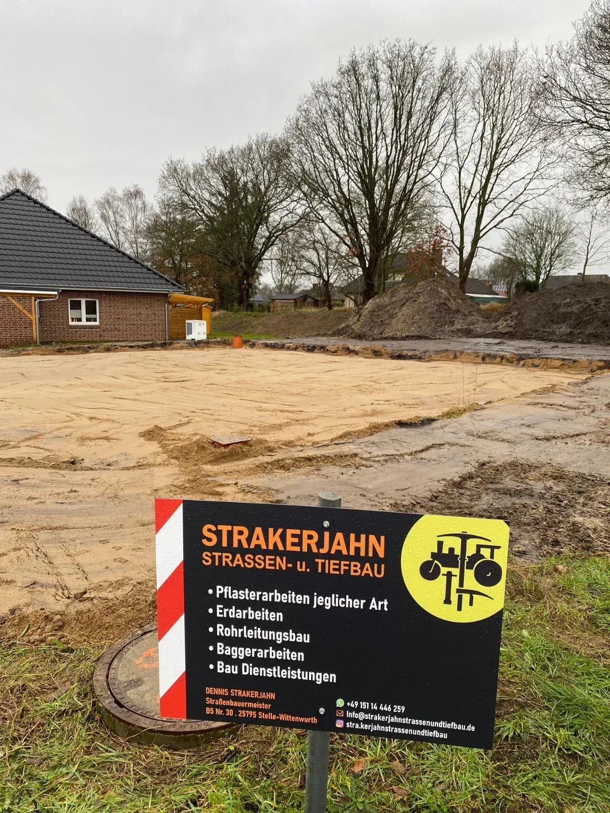 Schild für „Strakerjahn Straßen- u. Tiefbau“ auf einer Baustelle. Zu sehen sind eine Grafik mit Baumaschinen, ein Haus und Bäume im Hintergrund.