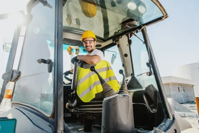 Ein Mann mit gelbem Schutzhelm und Sicherheitsweste lächelt aus einem Baustellenfahrzeug heraus.