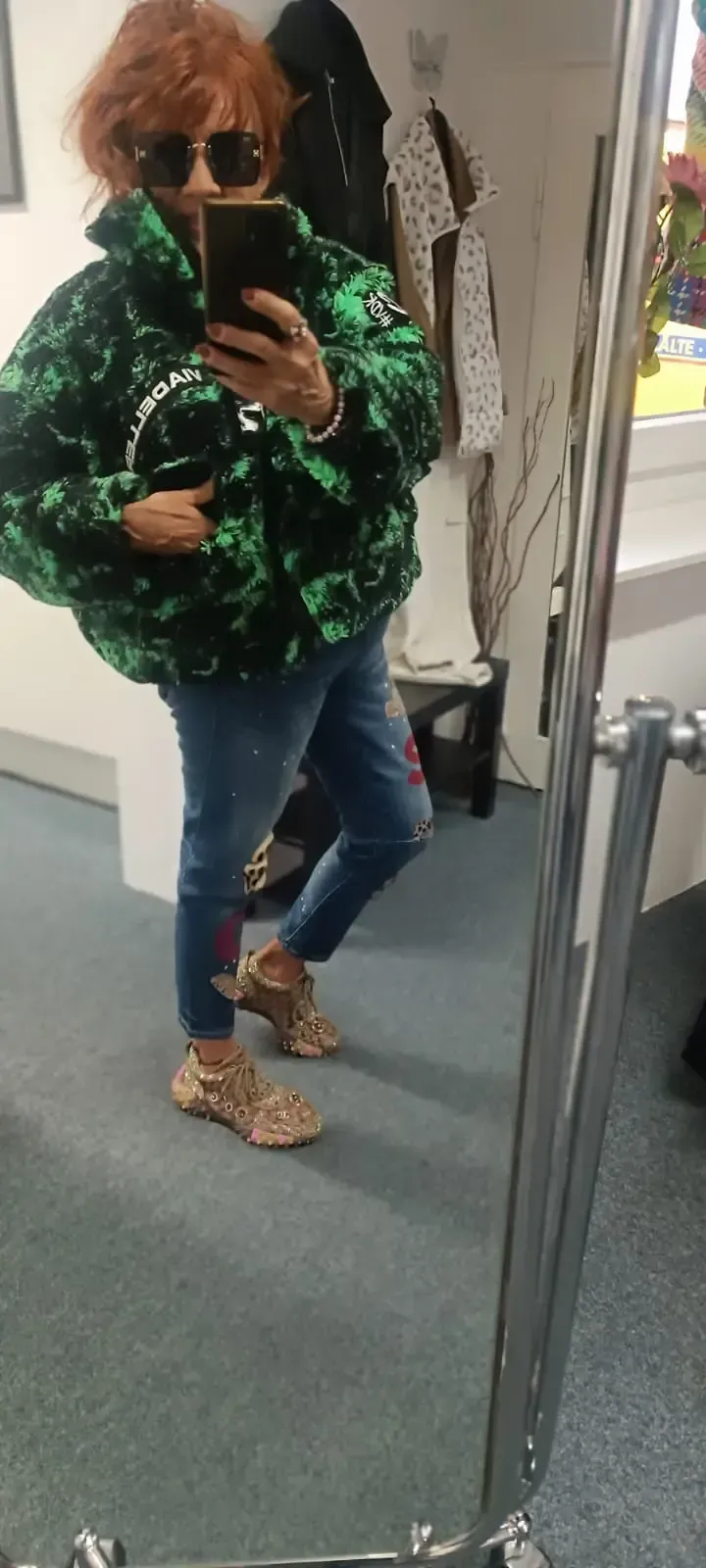 Eine Person macht ein Selfie vor dem Spiegel. Sie trägt einen grün gemusterten Pullover, blaue Jeans und Hausschuhe mit Leopardenmuster.