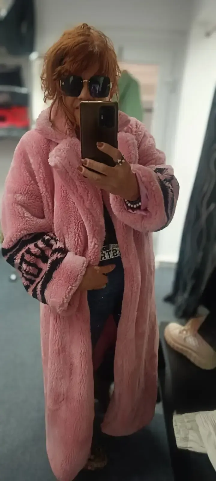 Spiegel-Selfie einer Person in einem rosa Pelzmantel und Sonnenbrille, die in einem Flur ein Handy hält