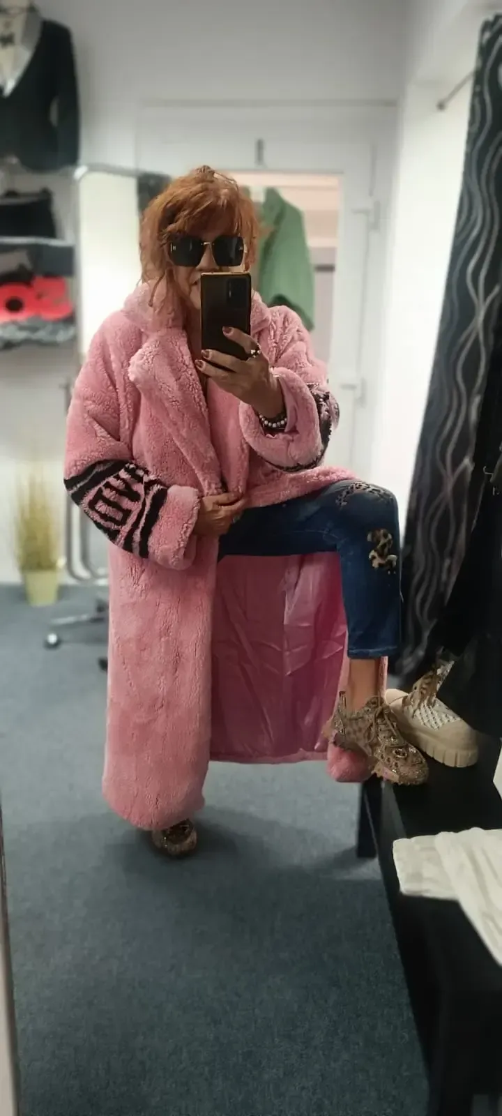 Eine Person, die in einem rosa Bademantel in einer Umkleidekabine sitzt und an einem Fuß einen Schuh trägt, macht ein Selfie im Spiegel.