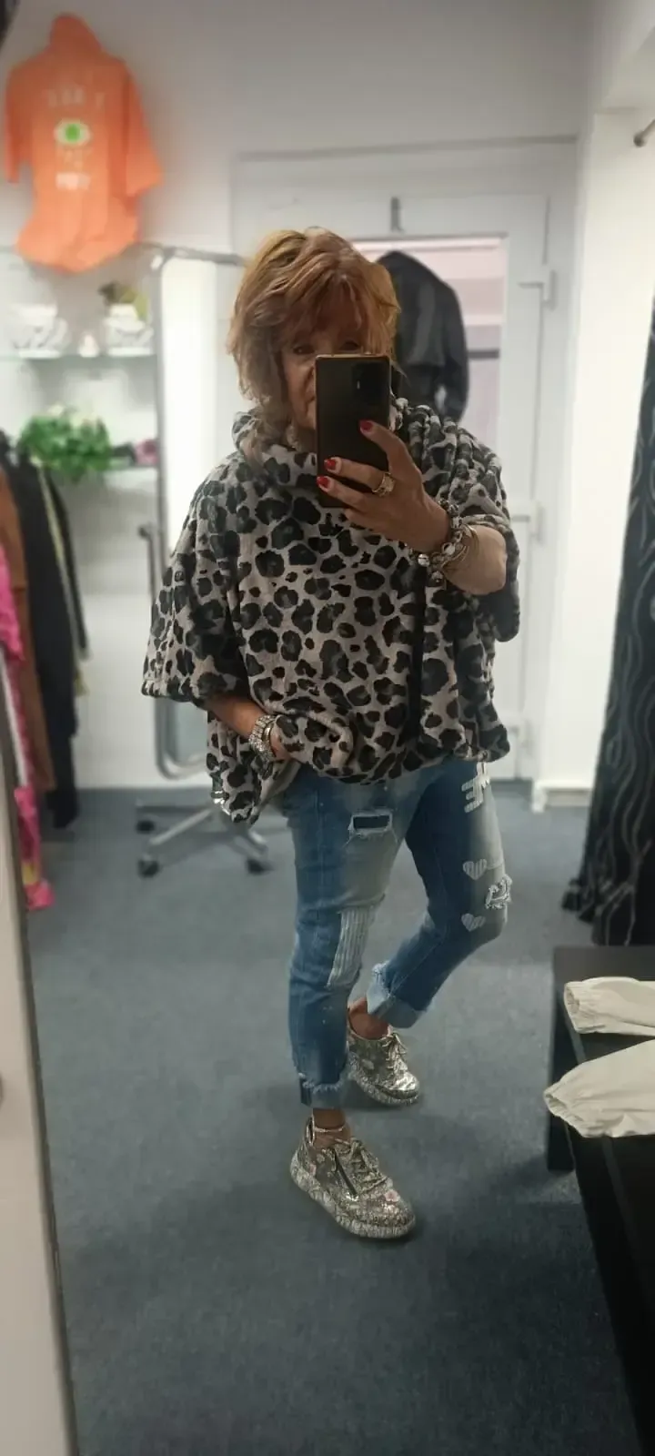 Eine Person macht in einer Umkleidekabine eines Bekleidungsgeschäfts ein Selfie vor dem Spiegel. Sie trägt ein Leopardenmuster-Top, zerrissene Jeans und Turnschuhe.