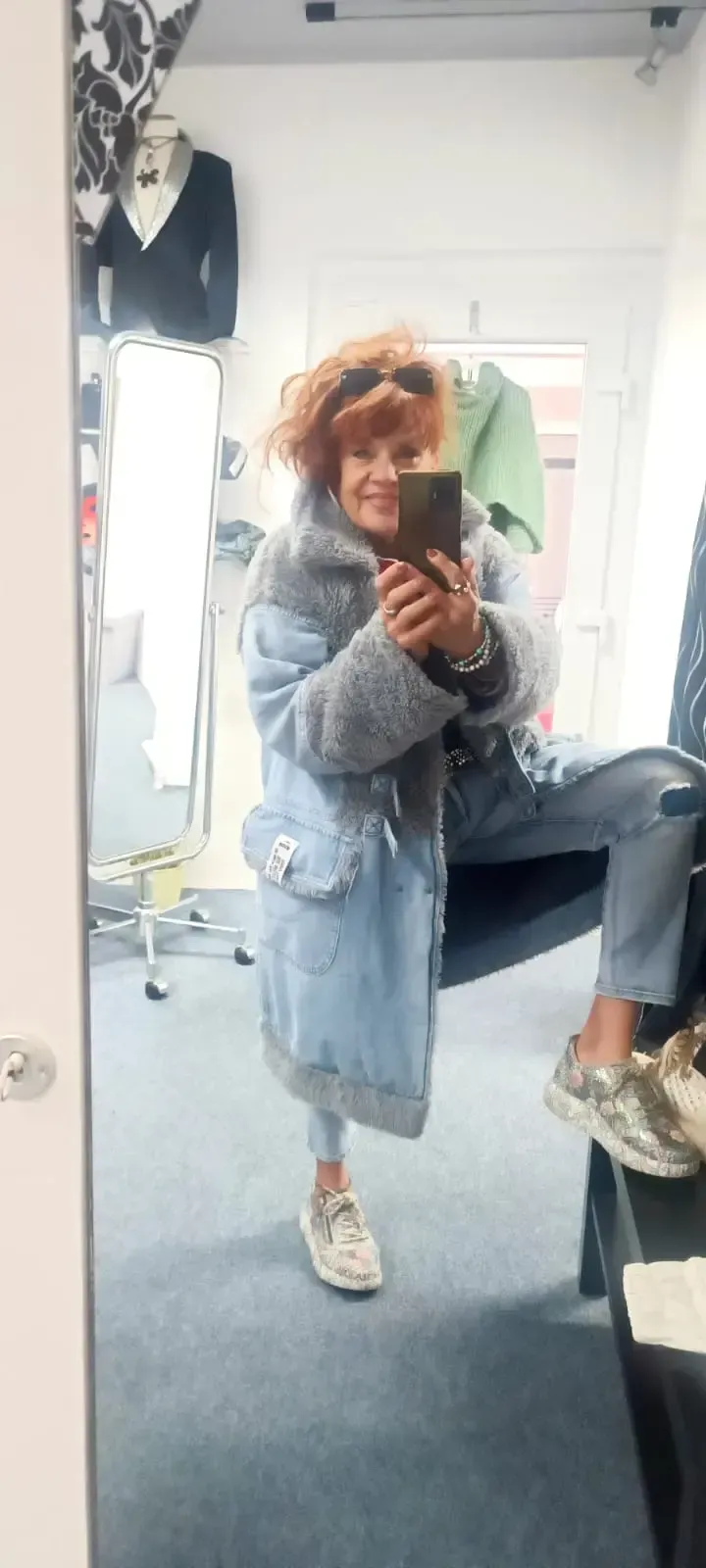 Eine Person, die in einem Umkleidekabinen-Spiegel hockt und ein Selfie macht, trägt einen hellblauen Mantel und weiße Turnschuhe.