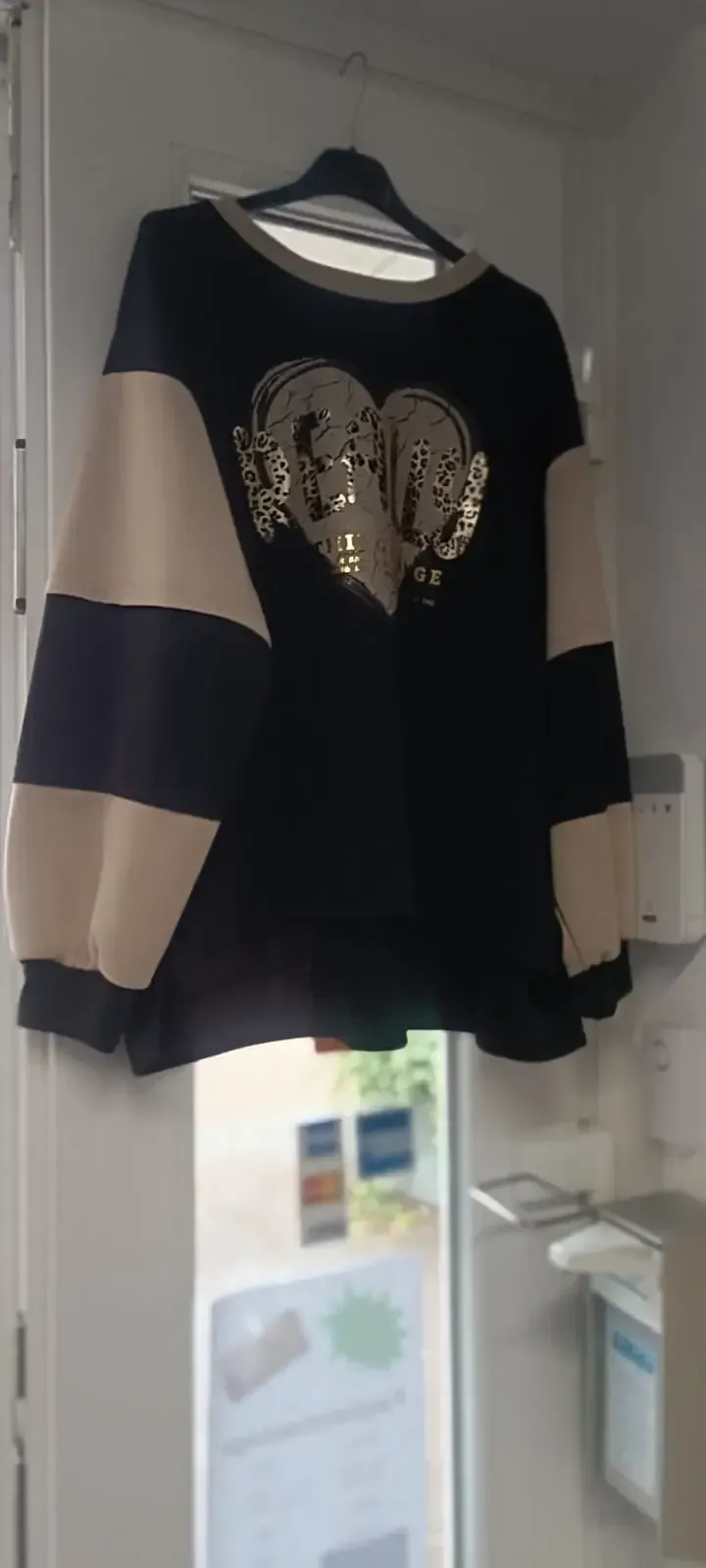 Schwarzer Pullover mit beigen Ärmeln und goldenem Elefantenmotiv hängt an einer Tür im Innenbereich.