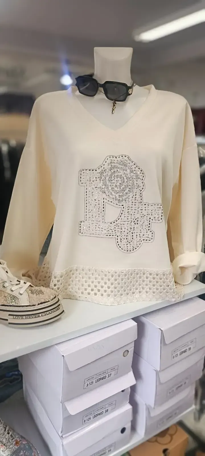 Ein cremefarbener Pullover hängt in einer Boutique an einer Schaufensterpuppe, darunter liegen gefaltete Kleidungsstücke in Regalen.