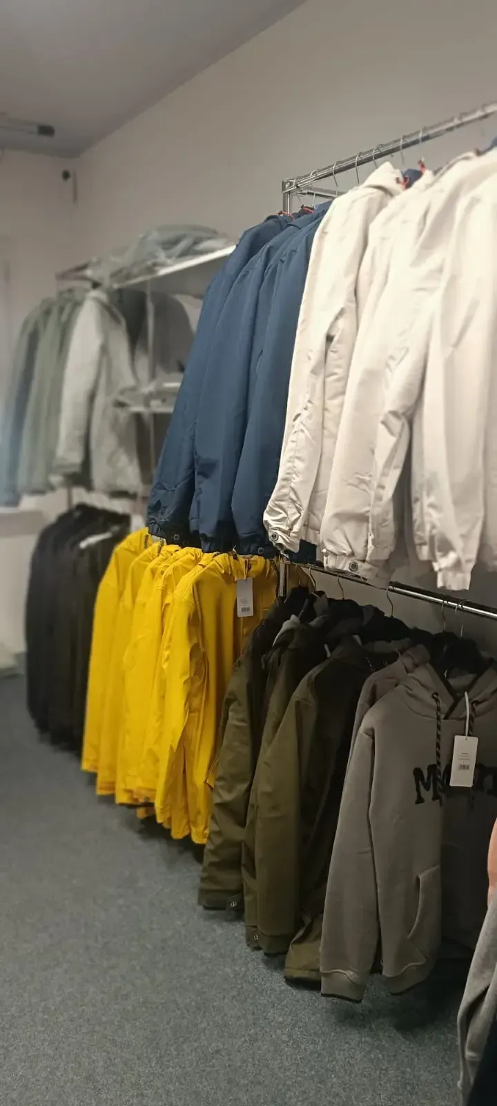 Kleiderständer mit Jacken und Hosen in Weiß, Blau, Gelb und Olivgrün, ausgestellt in einem Ladengang.