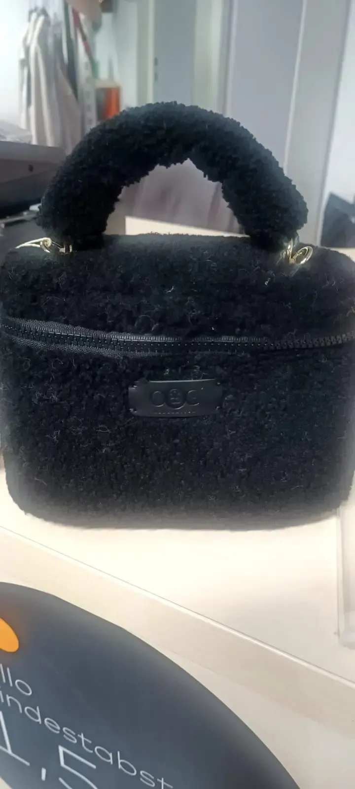 Schwarze, flauschige Handtasche mit abgerundetem Henkel auf einem Ausstellungsregal, dahinter eine weiße Schachtel.