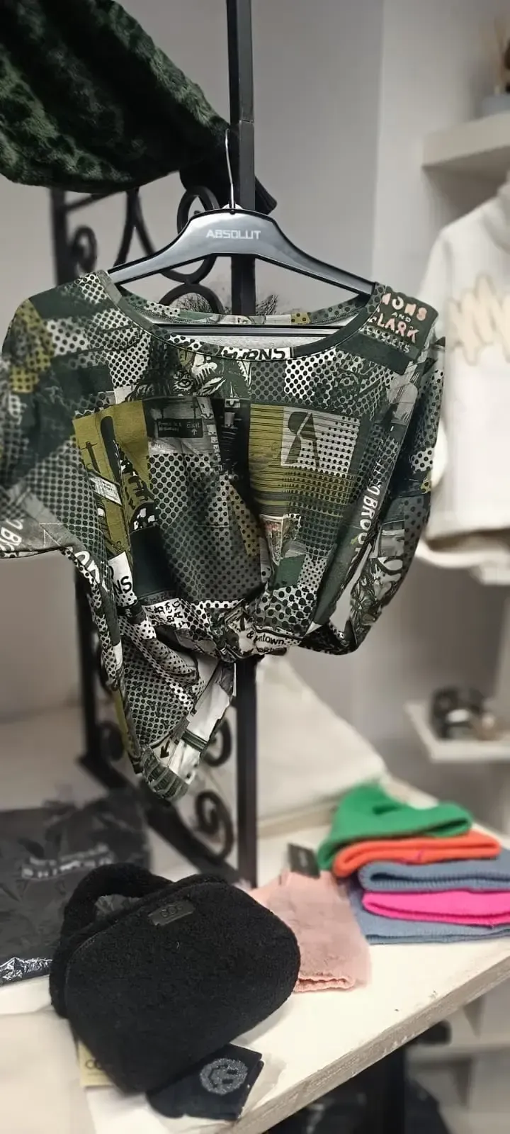 Ein Tarnhemd hängt neben gefalteter Kleidung an einem Kleiderständer, und eine schwarze Kappe liegt auf einem Regal.