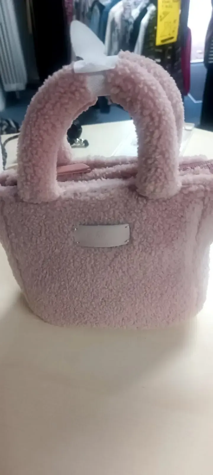 Eine pinkfarbene, flauschige Handtasche mit abgerundeten Henkeln steht auf einem Tisch; im Hintergrund sind Kleiderständer verschwommen zu erkennen.
