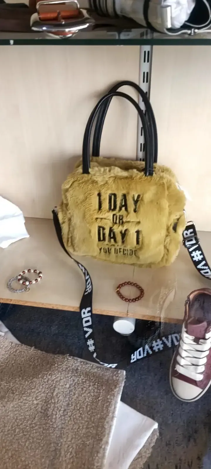 Gelbe Handtasche mit schwarzen Henkeln und Aufdruck, ausgestellt auf einem Regal neben Turnschuhen.