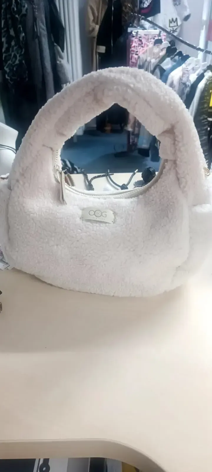 Eine kleine weiße Plüschhandtasche mit abgerundetem Henkel steht auf einem Tisch in einem Schaufenster.