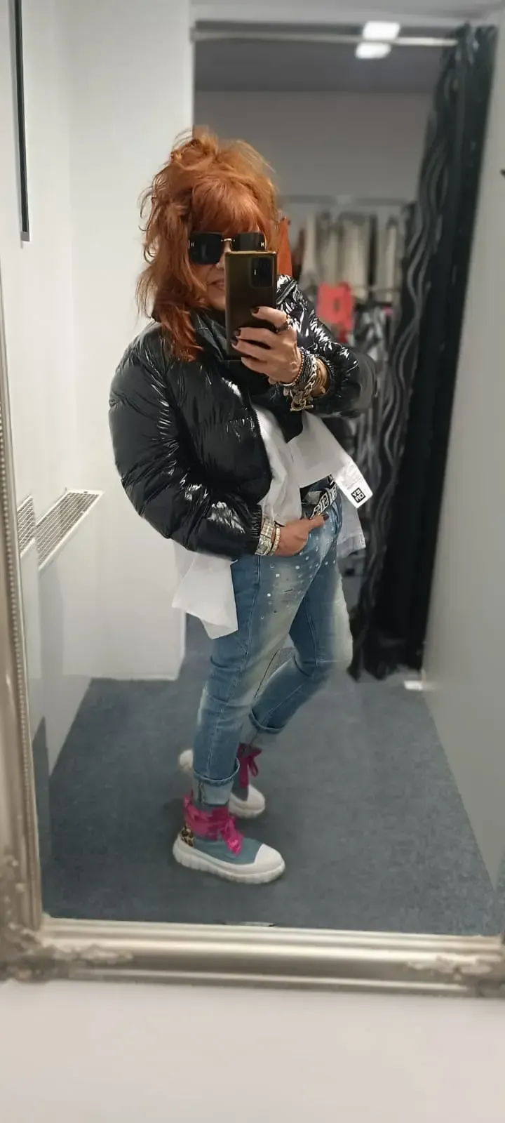 Eine Person, die in einer Umkleidekabine ein Spiegel-Selfie macht, trägt eine schwarze Jacke, Jeans und pinkfarbene Turnschuhe.
