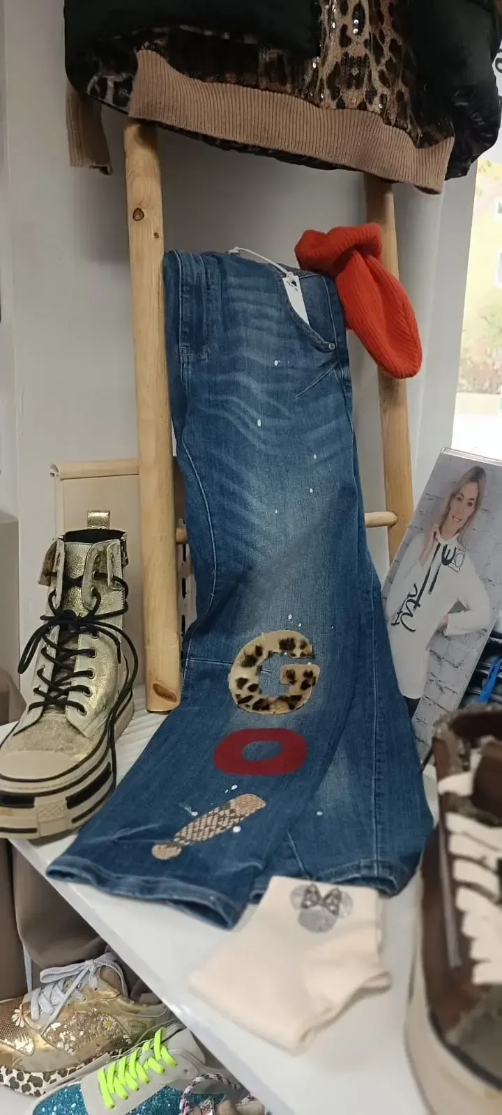 Eine blaue, zerrissene Jeans, vertikal ausgestellt mit einem roten Handschuh, umgeben von Schuhen und Kleidung in einem Schaufenster