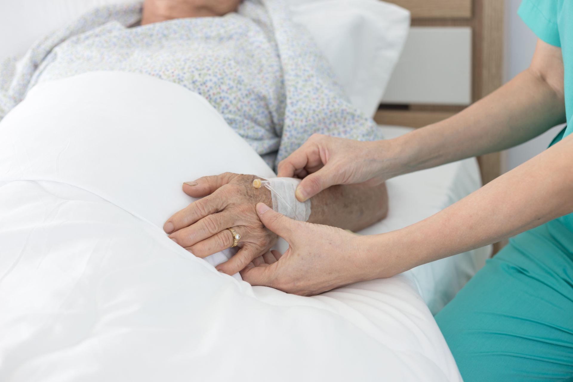 Une soignante tient la main d'un patient alité dans un hôpital aux draps blancs.