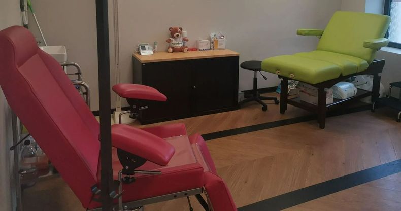Couloir étroit bordé de chaises colorées le long des deux murs et se terminant par une double porte en bois fermée.