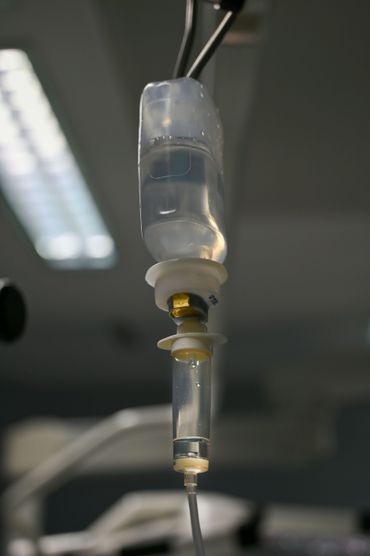 Flacon de perfusion intraveineuse au premier plan, chambre d'hôpital floue et patient alité en arrière-plan.