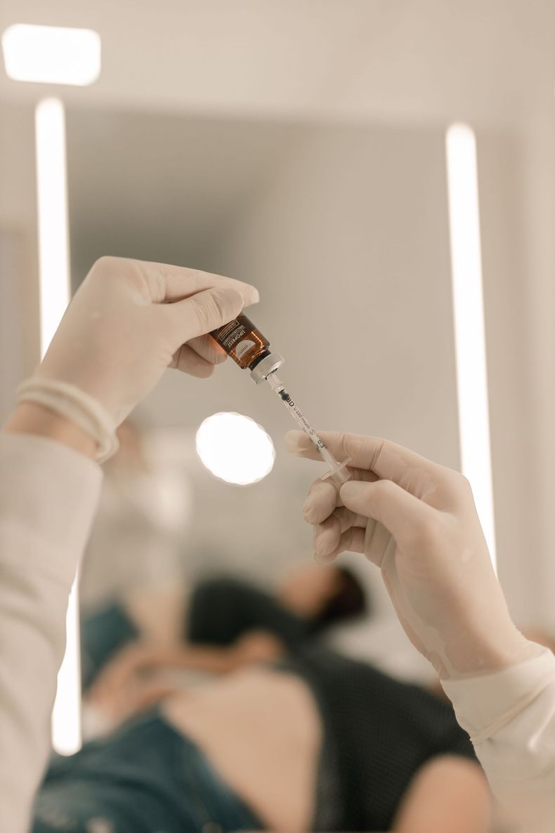 Des mains gantées préparent un flacon d'injection dans un environnement clinique, avec un patient flou en arrière-plan.