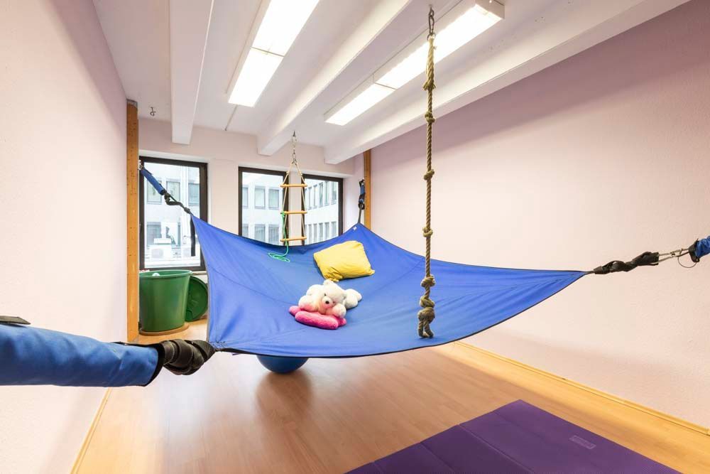 Ein Kind liegt in einer blauen Hängematte in einem hellen Zimmer und hält ein gelbes Kissen und ein Stofftier.