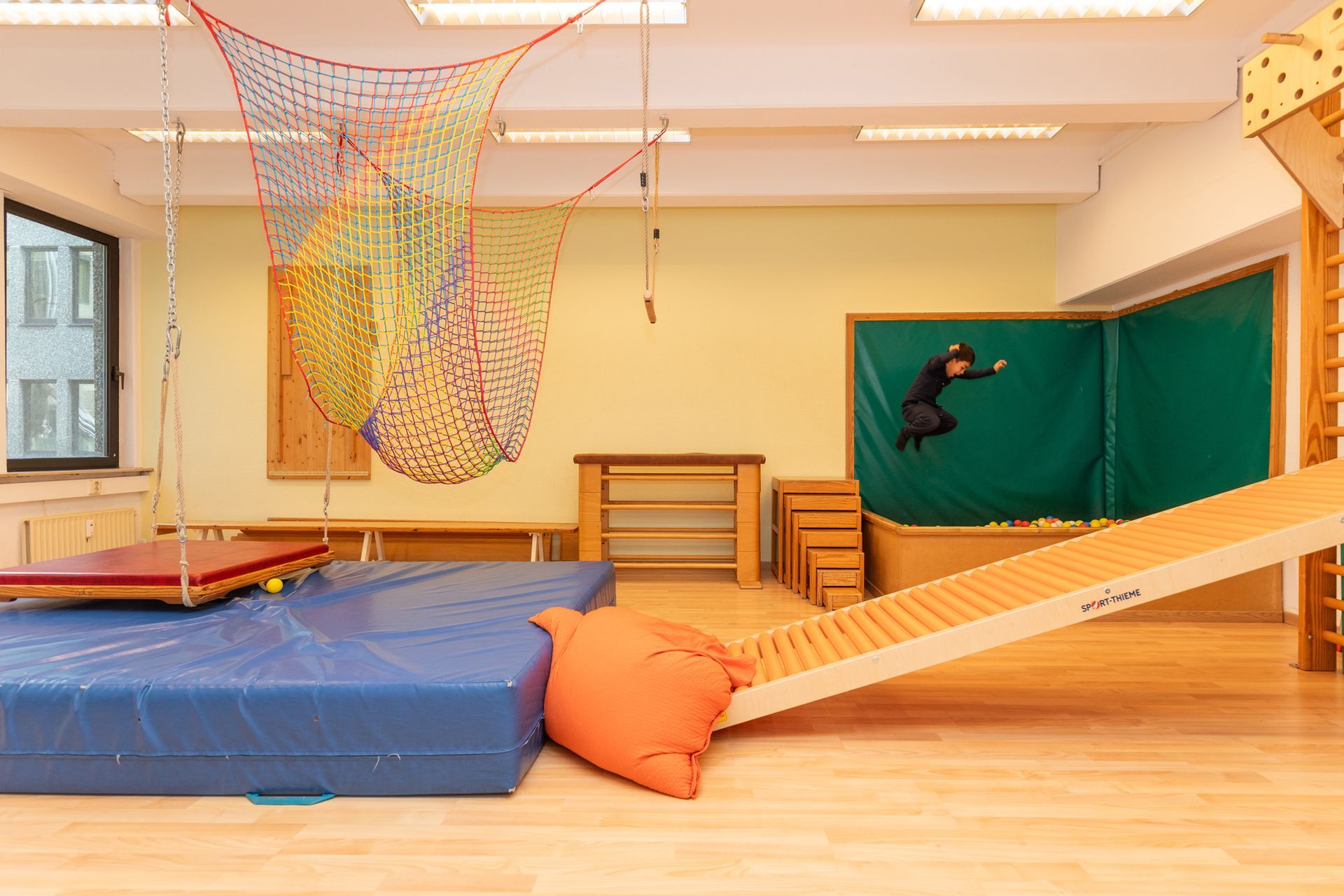 Indoor-Turnhalle mit blauen Matten, Seilnetz, Rampen und einem Kind, das an einer grünen Wandwand hochklettert.