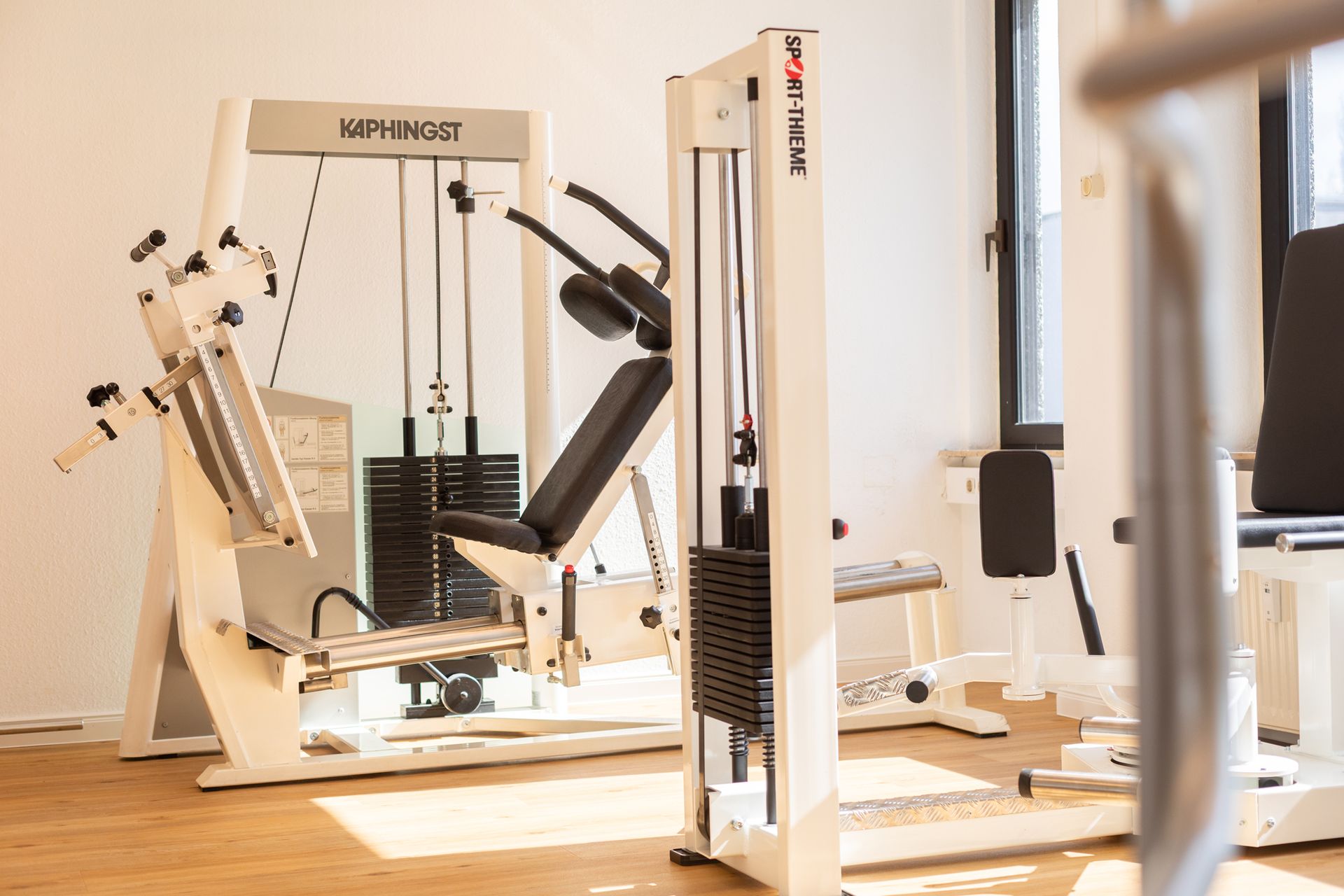 Fitnessgeräte in einem hellen Studio, mit weißen Krafttrainingsgeräten und schwarzen Sitzen.