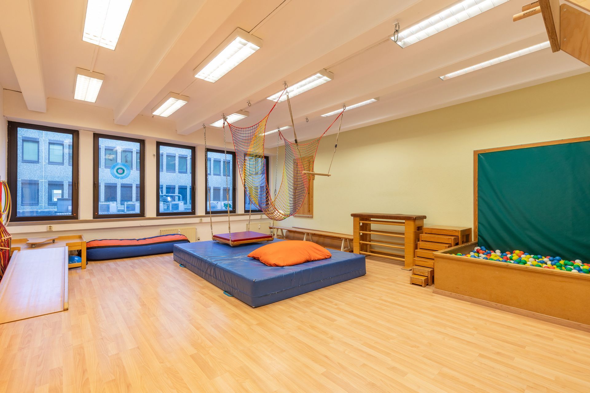 Helles Klassenzimmer mit blauen Matten, Fenstern, Holzboden und einer grünen Tafel an der Vorderseite