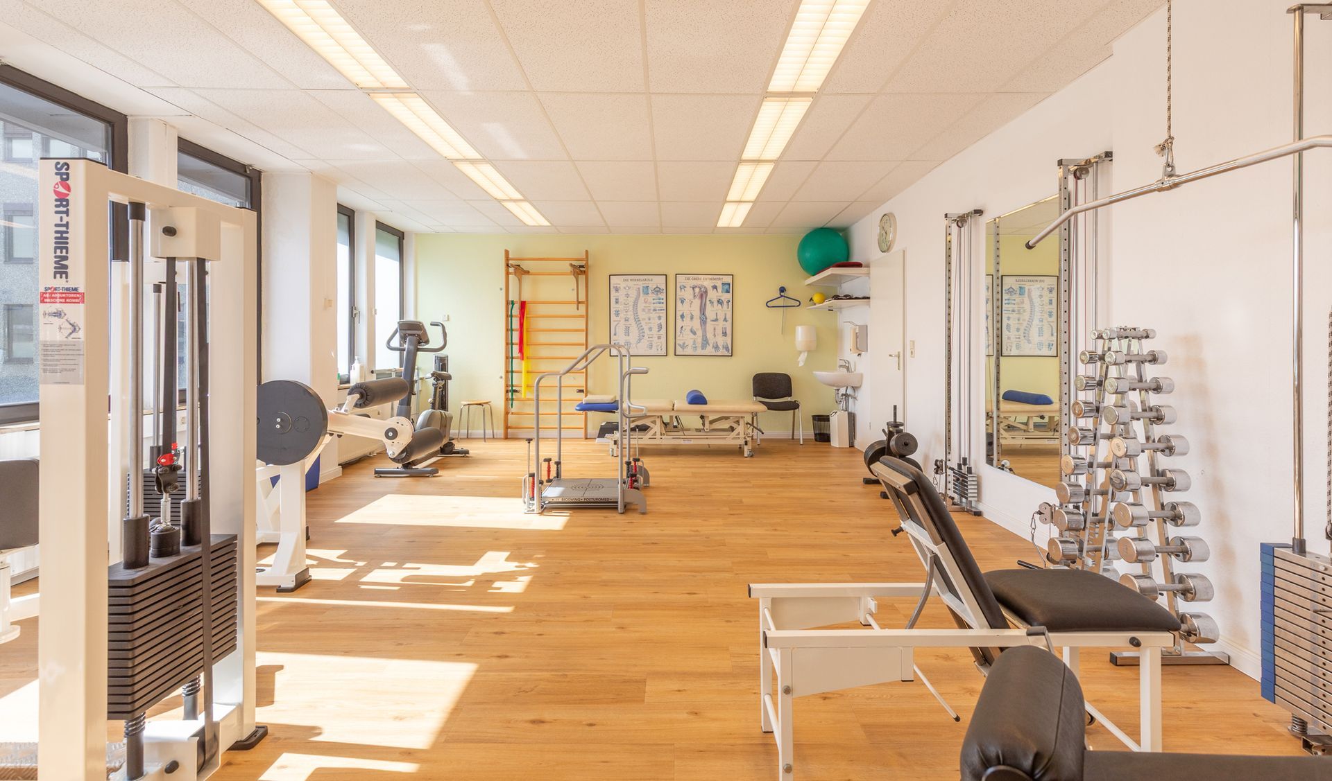 Heller Physiotherapieraum mit Holzboden, Trainingsgeräten, Spiegeln und Wandtafeln.