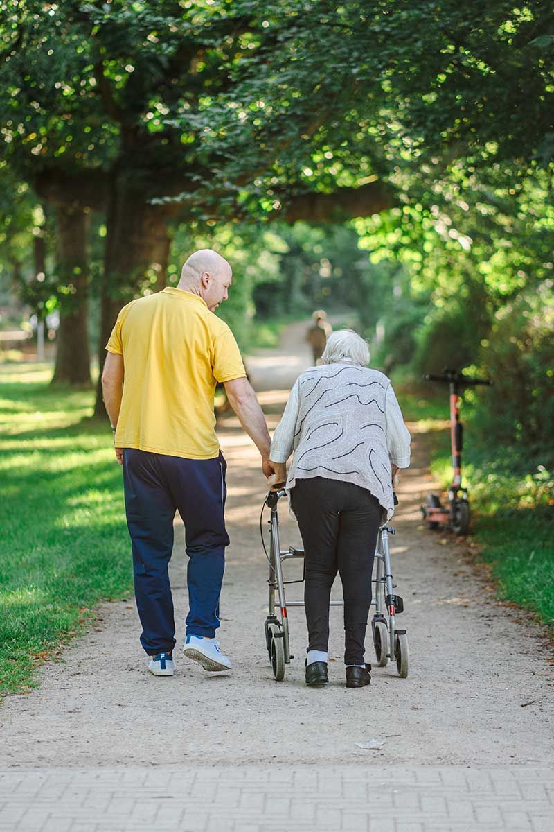 Zwei Personen gehen auf einem von Bäumen gesäumten Weg entlang, eine führt die andere mit einem Rollator.