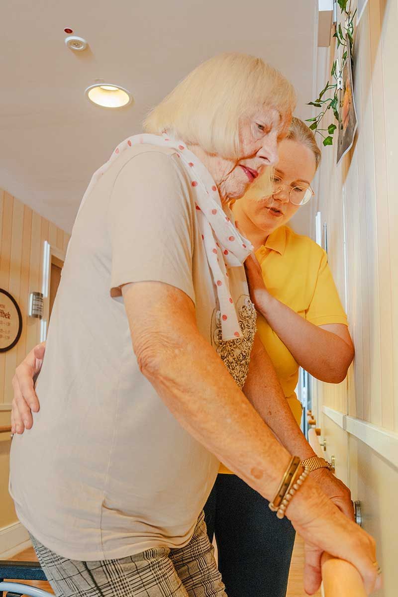 Zwei Personen umarmen sich drinnen an einem Fenster, eine in einem hellen Hemd, die andere in einem gelben Oberteil.