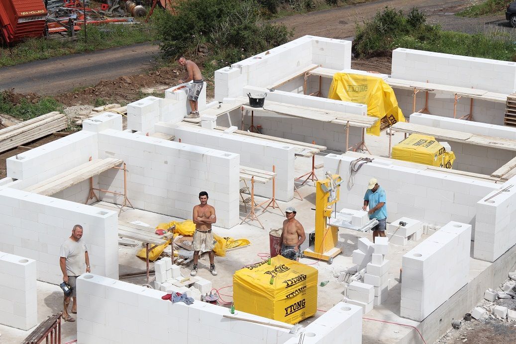 Des ouvriers assemblent la structure d'une maison en mousse blanche sur un chantier de construction, à l'aide d'équipements et de bâches jaunes.