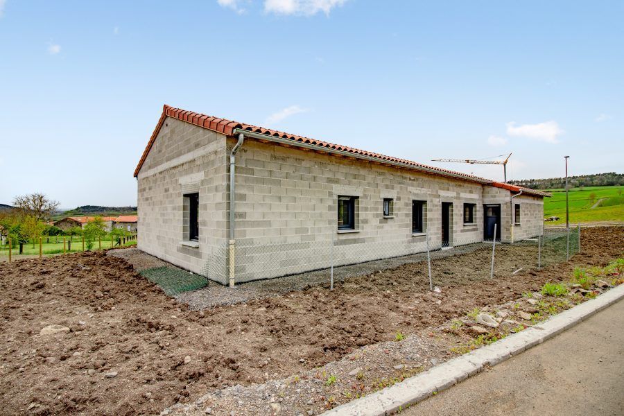Maison neuve en briques grises sur un terrain en terre battue, avec un toit rouge, dans une zone rurale.