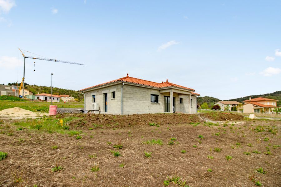 Maison de plain-pied en construction sur un terrain vague, avec une grue et un ciel bleu azur.