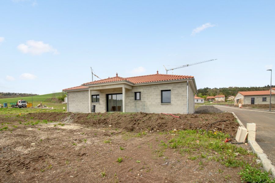 Maison de plain-pied en construction sur un terrain en terre battue, avec un toit de tuiles rouges et des murs extérieurs inachevés.