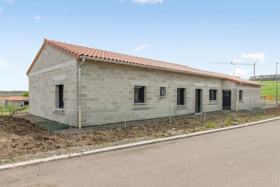 Maison neuve de plain-pied en briques grises avec un toit de tuiles rouges, située au bord d'une route pavée.