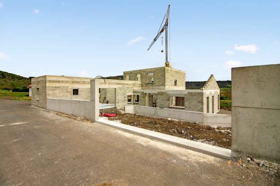 Petite maison en béton en construction en bord de route, avec une éolienne sur fond de ciel bleu azur.