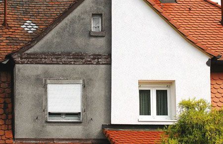 Deux façades de maisons mitoyennes, l'une grise et l'autre blanche, avec des toits et des fenêtres en tuiles rouges.